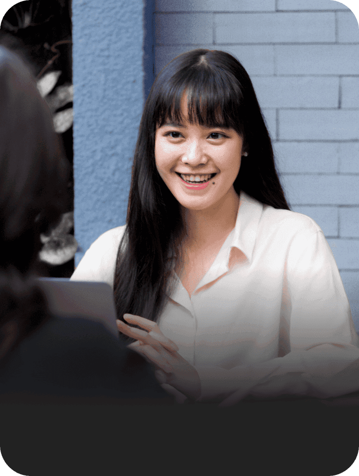 A woman in a white shirt smiles while holding a laptop during a meeting, engaging with the person across from her.