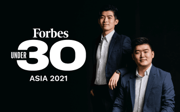 Topremit co-founders pose for Forbes 30 Under 30 in 2021, with Hermanto Wie seated in a dark gray suit and smiling, and Henry Wirawan standing in a black suit.