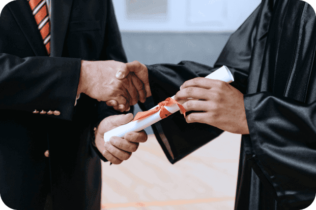 Two people wearing graduation robes shake hands while handing over a rolled-up paper.