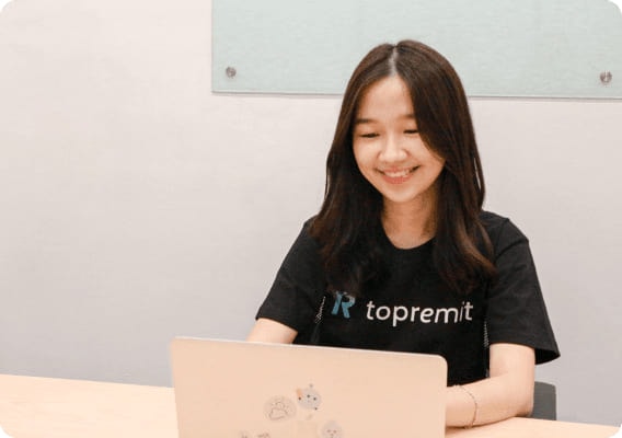 黒いTopremitのTシャツを着た社員が、ノートパソコンで作業しながら笑顔を見せています。